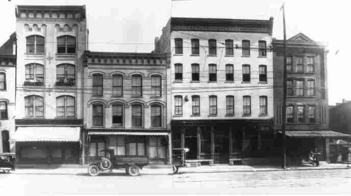 Historic black-and-white photo of a row of downtown storefront buildings, an early delivery truck parked out front